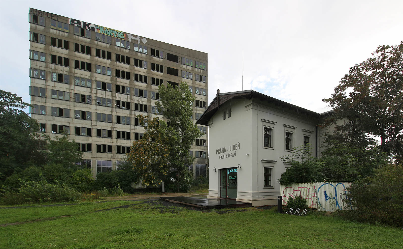 Kolejiště stanice Praha-Liběň dolní nádraží se nacházelo severně od staniční budovy (na snímku blíže k fotografovi), mezi histroickou budovou a současným betonovým monstrem se nacházelo několik kolejí vleček a rozsáhlý areál skladů, ze kterých nezůstalo nic. Koleje odsud ale tak úplně nezmizely: přímo pod cípem centra Nová Palmovka prochází od roku 1990 tunely metra B.