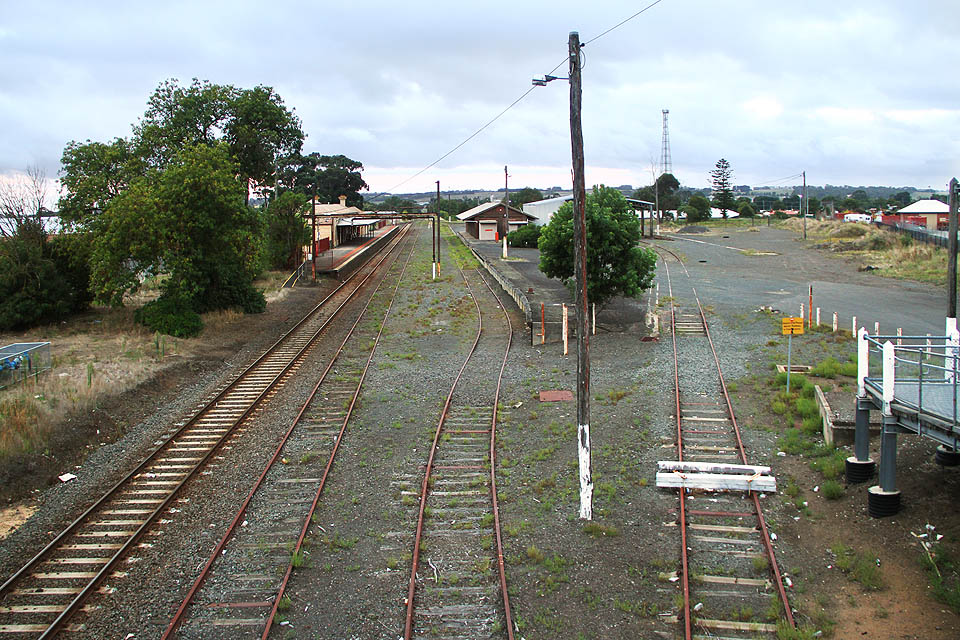 Čtyři koleje železniční stanice Colac. Vlevo dopravní kolej s krytým nástupištěm u výpravní budovy, vpravo bývalá dopravní kolej č. 2 a manipulační koleje u rampy a na překladišti se zrušenou úzkokolejnou drážkou. Na místě úzkokolejiště se rozkládá šedivá štěrková plocha.