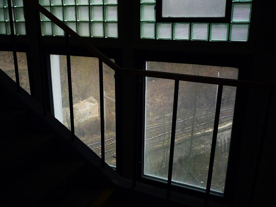 Pohled z jedné opuštěné budovy na druhou. Z mrakodrapu uvnitř továrny je vidět vlaky projíždějící skalnatým údolím okolo strážního domku, ke kterému nikdy nevedla cesta.