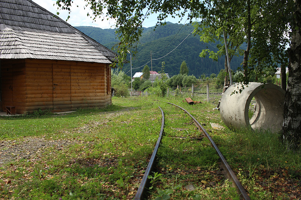 Koleje prochází podél plotu skanzenu za stěnami roubených lidových chalup.