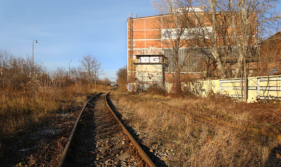 V oblouku se vlečková kolej (na snímku) a staniční koleje (pod stožáry vlevo) poněkud vzdalují, odděleny pásem bujného křoví. Před bílými vraty uprostřed snímku se nachází čtvrtá brána na vlečce.