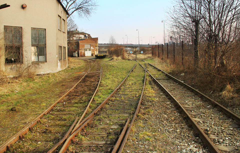 Střídmý pohled za pátou bránu. Dvě koleje vlečky pokračují hlouběji do areálu, jedna se vrací skrz další bránu ven, aby se na chvíli spojila se staniční kolejí před definitivním zvlečkovatěním bývalé dráhy.