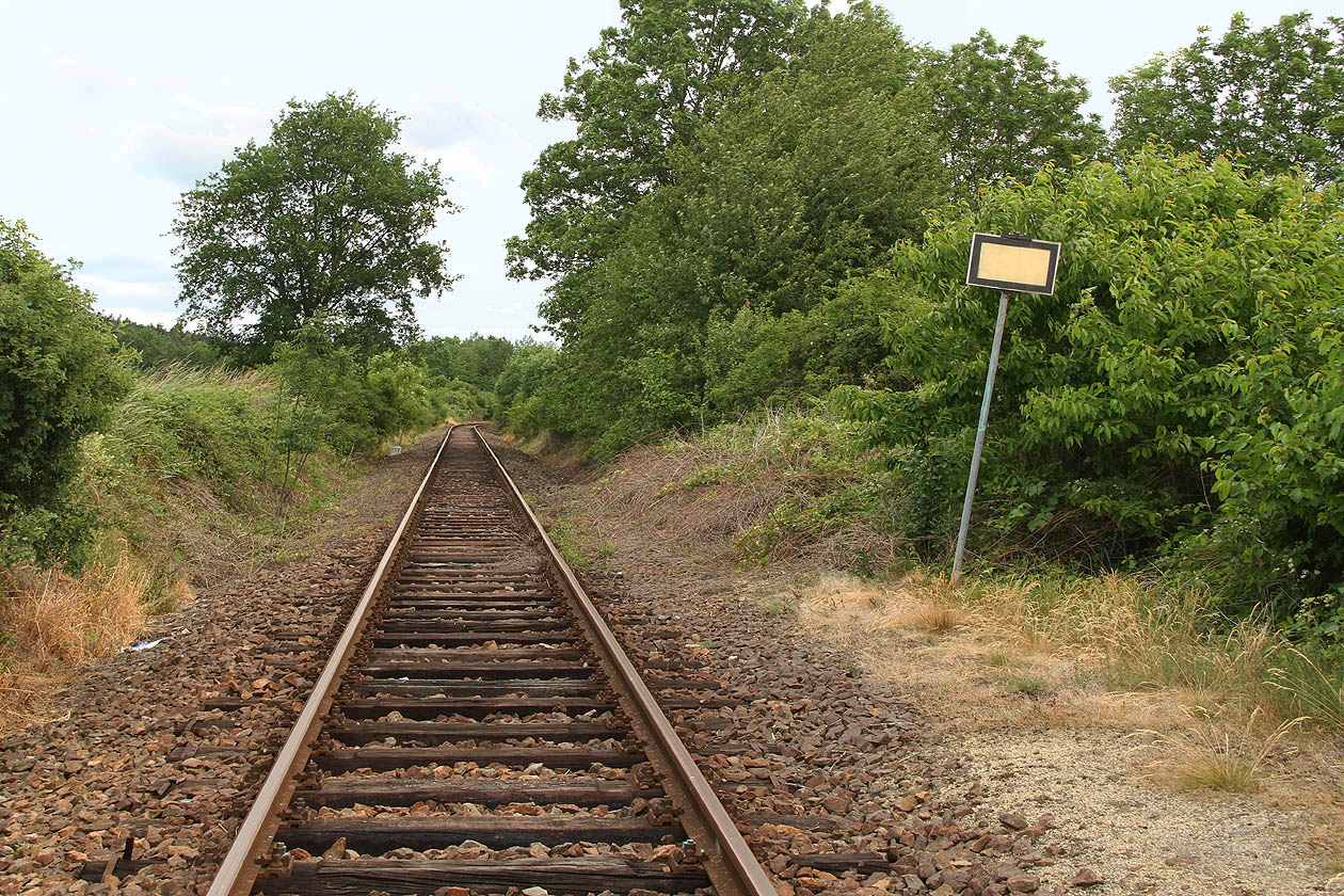 Cedulka označující konec nástupiště dlí daleko za koncem nástupiště v suché trávě a křoví.