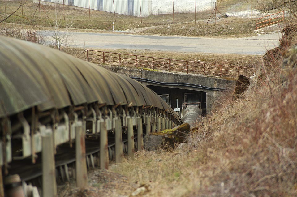 Ze zásobníku kameniva pod silnicí vedou oba transportní pásy pod zemí. Vypadají jako obrovská lesklá stonožka, která vylézá ze své škvíry pod kamenem. Ze zásobníku kameniva pod silnicí vedou oba transportní pásy pod zemí. Vypadají jako obrovská lesklá stonožka, která vylézá ze své škvíry pod kamenem.
