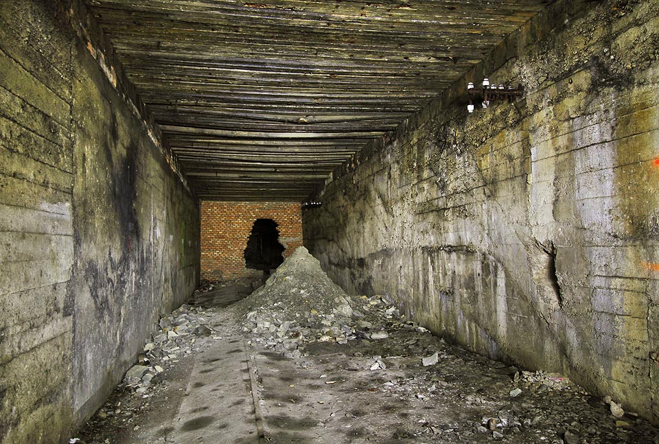 Vstupní část kmenové štoly D je v současnosti jediným alespoň částečně bezpečným místem v přístupné části podzemní továrny. Přesto zde vznikají menší závaly. V levé části je dobře vidět deformovaná podlaha s drážkou rozchodu 600 mm nadzdvižená nabobtnalými slínovci.