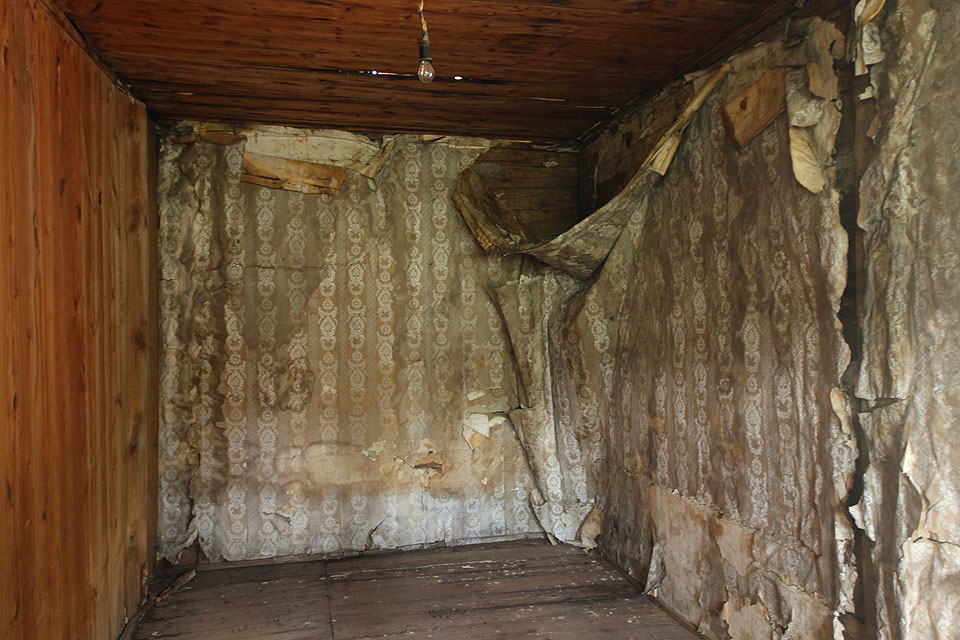 Ložnice či dětský pokoj se zpuchřelými tapetami odhaluje prohnilým otvorem ve stropě nedokonalosti prejzové střechy.