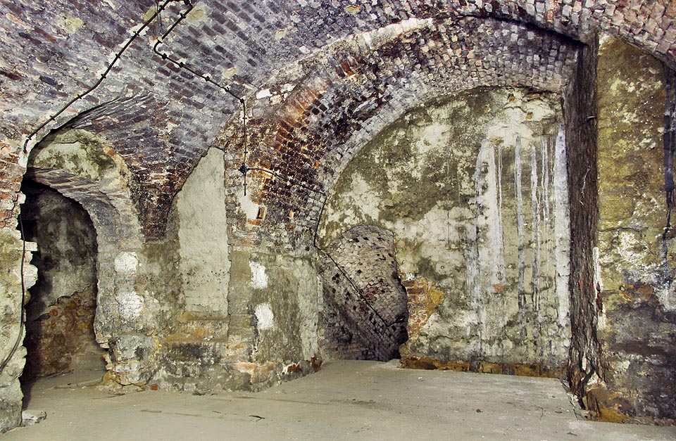 Centrální sklep za sálem s pasy: románské kamenné zdivo, zazděné schodiště do vyšších pater a barokní konstrukce masivních křížových kleneb. Další schodiště klesá do druhého podzemního poschodí, kde končí zazdívkou.