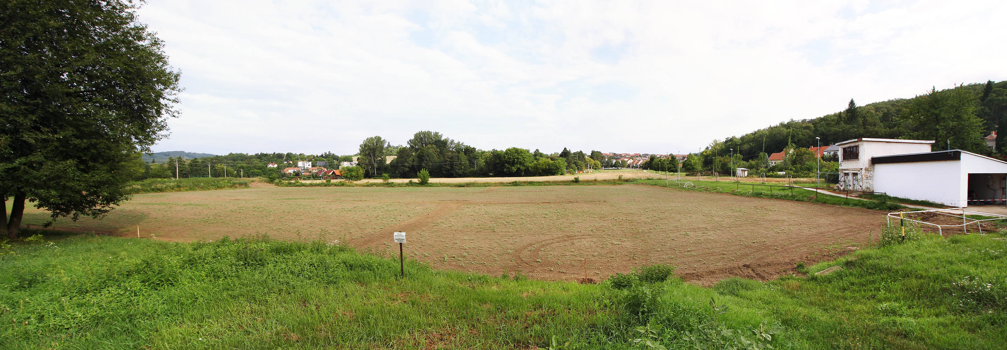 Tak vypadalo místo zamýšlené ke stavbě úpravny vody v roce 2017. Nivu nad soutokem Svratky s Mniším potokem pokrývá zorané fotbalové hřiště a pole. V popředí na fotce se měly nacházet rozlehlé skládky písku pro pozvolné filtry, samotné filtry by pak stály na rozhraní žlutého pole a lesa v dálce. Na místě domů v pozadí by stála budova s usazovacími nádržemi a hrubocezy, za bílým domem vpravo pak hlavní vodojem a strojovna čerpací stanice.