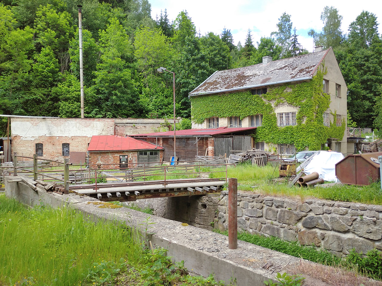 Pohled, který se naskytne kolemj(e)doucím ze silnice vedoucí po dně údolí. Před svým zasypáním vedl za hlavní budovou pily, obrůstající břečťanem, vodní náhon, který zásoboval továrnu energií.