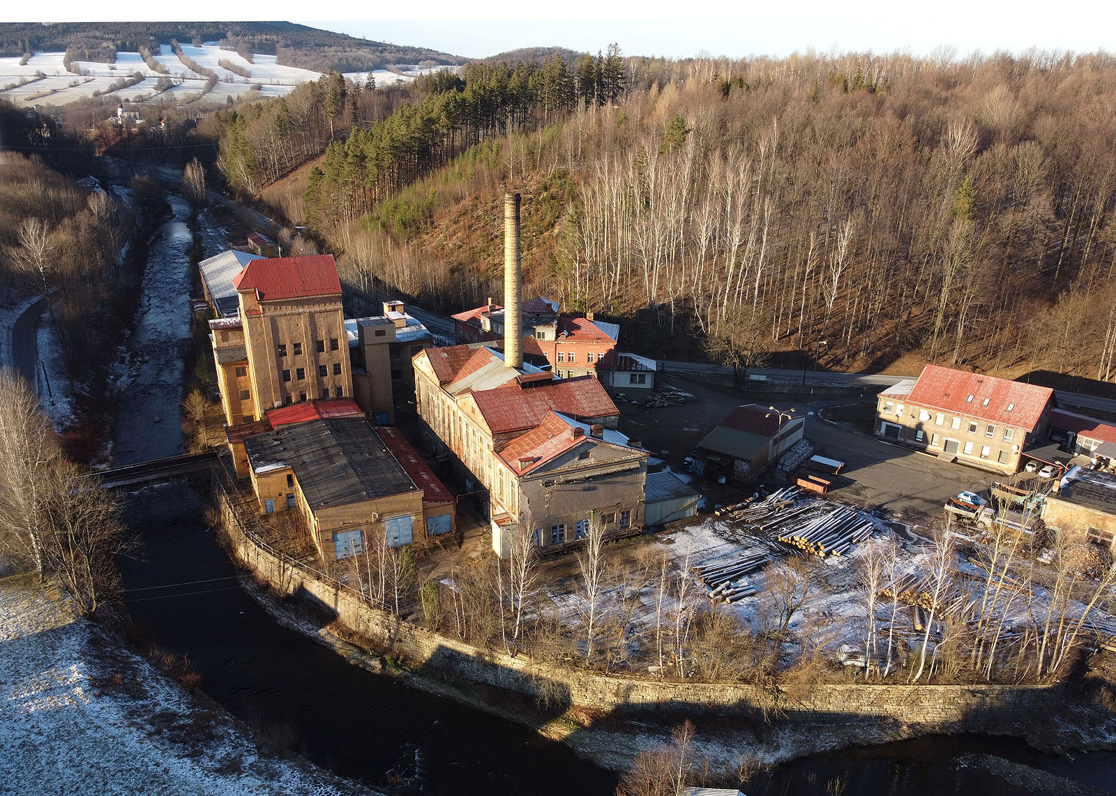 Pohled na bývalou papírnu, později továrnu na zemědělské přívěsy čtyři dny před koncem roku 2020. Stísněné údolí neumožňovlo umístit skládku dřeva dole u řeky a proto musela být vybudována vzadu nad továrnou z pohledu fotografa.