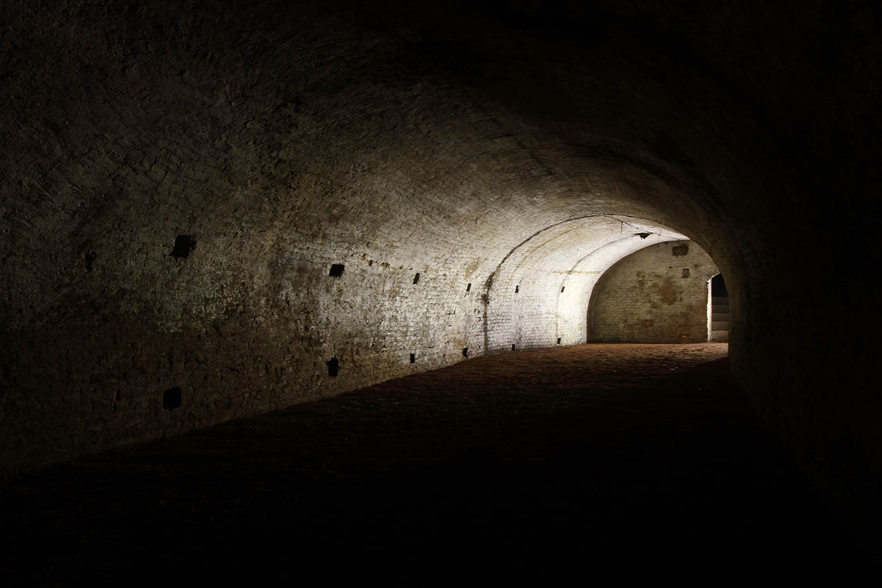 Od schodiště (na fotce vpravo) se místnost zatáčí táhlým obloukem doleva. Na obou stranách jsou ve stěnách v pravidelných rozestupech zahloubené otvory, jež snad sloužily k upevnění konstrukce regálů či polic na skladování zboží.