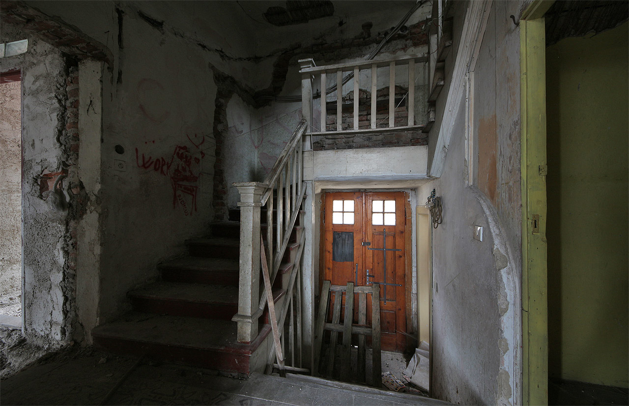 Hlavní vstup a jemně zdobné dřevěné schodiště. Snaha o dlouholetou a nikdy nedokončenou rekonstrukci je patrná na každém kroku. Ze stěn domu sálá osamění a smutek.